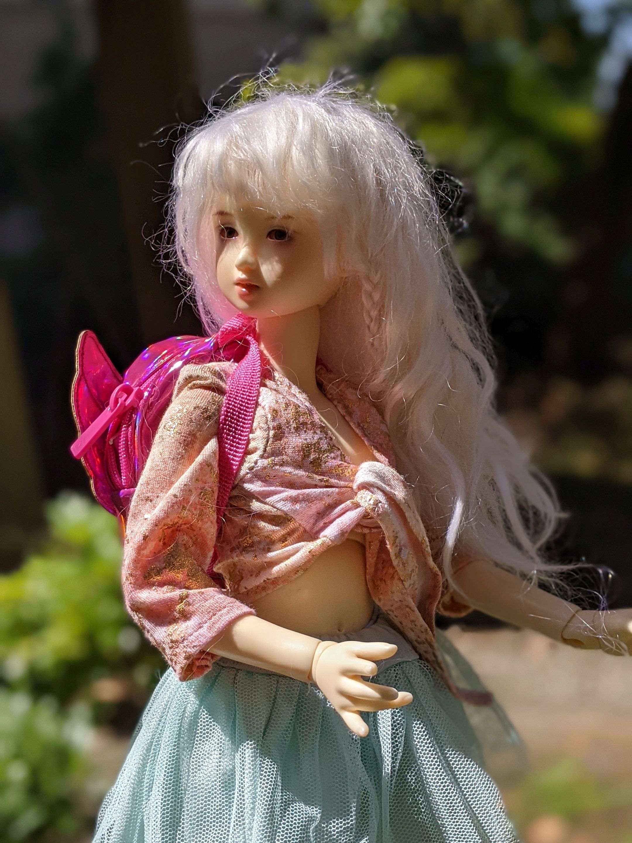 a photo of a girl doll looking up off to the left. she is white and has white hair. she has a pink top tied around her waist and a pink backpack.