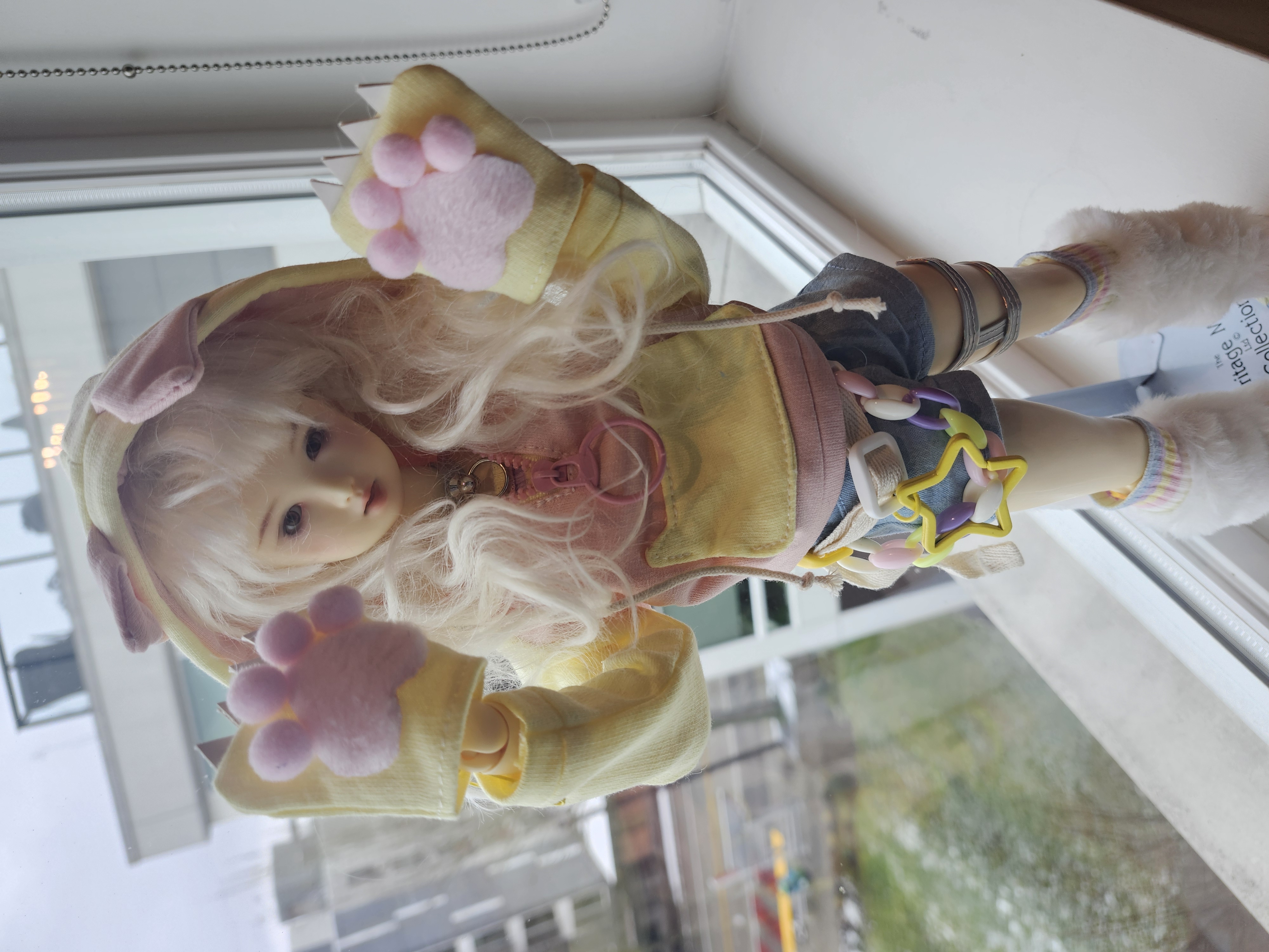 a photo of a girl doll looking up up to the viewer with her arms up in the air in a pouncing pose. she is white and has white hair. she is wearing a yellow outfit and has animal paw gloves.
