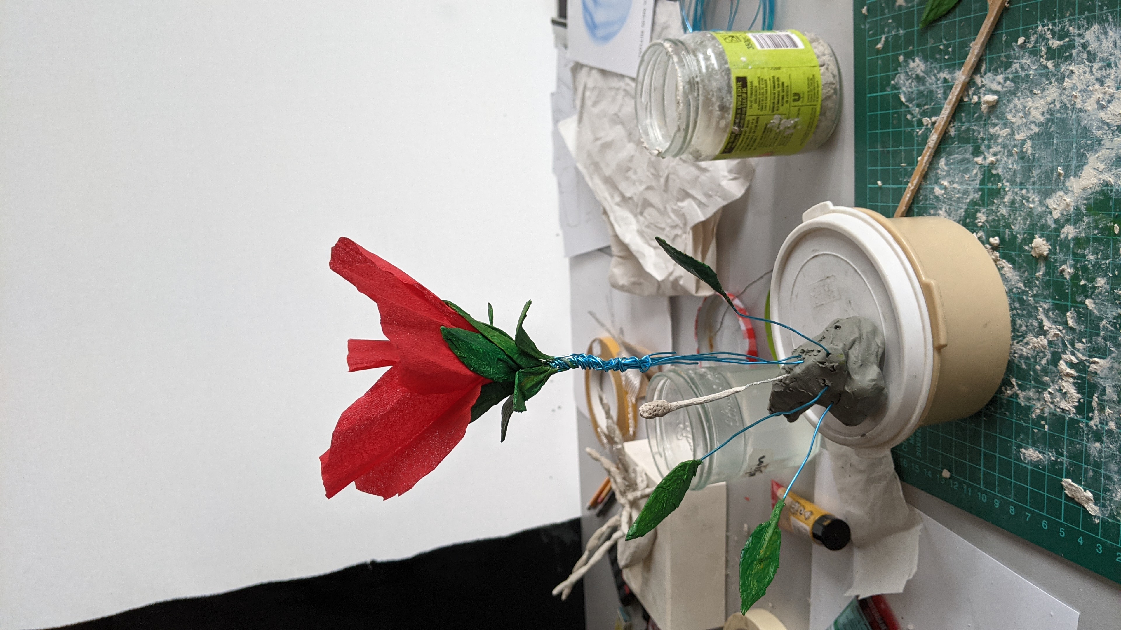 a photograph of a table that has a sculpture of a red hibiscus flower that is in the process of being worked on. it loooks like it's handmade from paper, wire, and paint. behind it, in the background sits a scultpure of a spider that is also under construction and is not painted yet. the table is messy and has supplies like paint, paper, glue, and wire strewn across it.